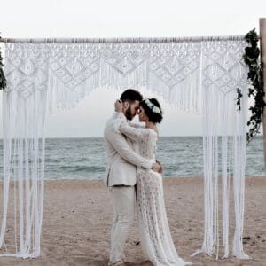 Couple embracing under a white macramé arch by the beach.