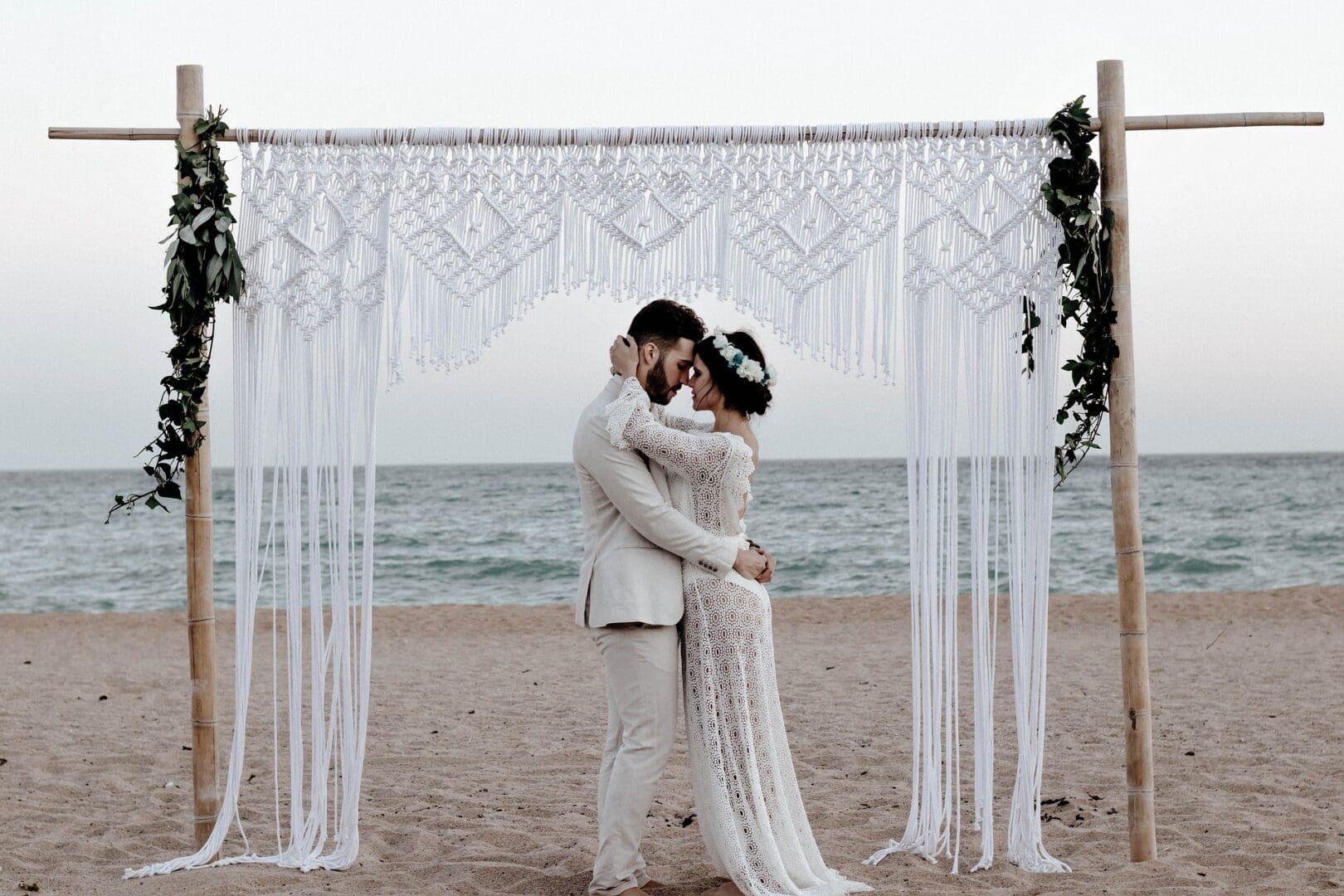 Couple embracing under a white macramé arch by the beach.