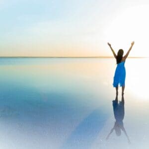 A person with arms raised stands on a calm, reflective water surface at sunset.
