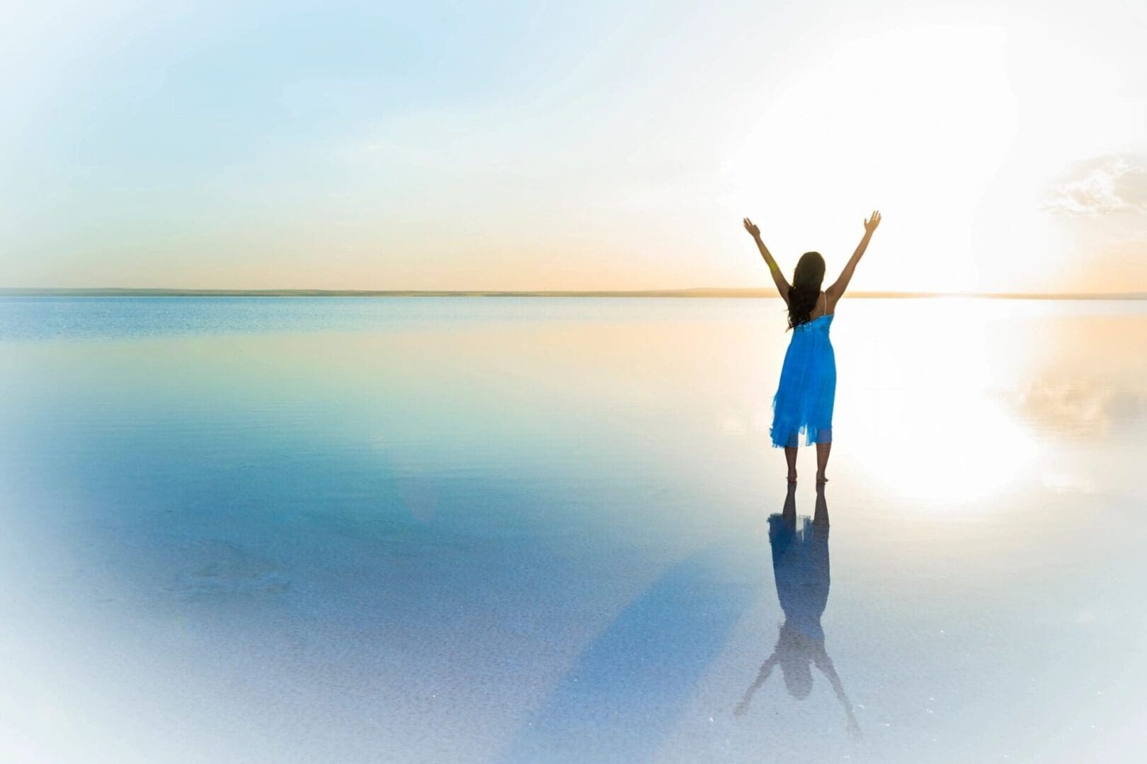 A person with arms raised stands on a calm, reflective water surface at sunset.