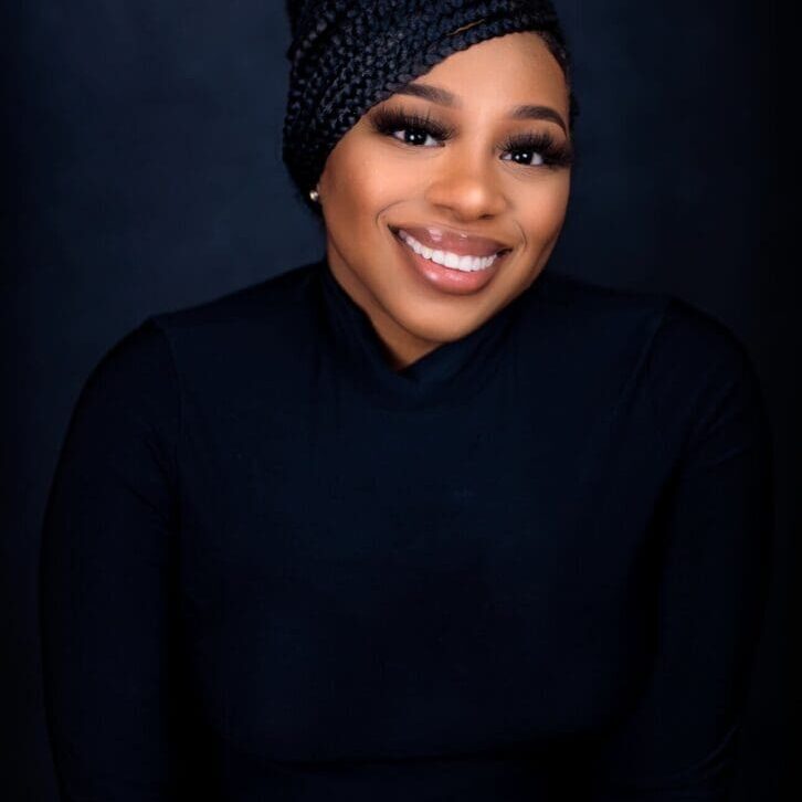 A smiling woman wearing a dark headwrap and black top against a black background.